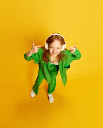 Happy girl, teenager with red hair wearing stylish green costume wearing wireless headphones over yellow studio background. Top view. Concept of youth, fashion, music, lifestyle, hobby, emotions, ad