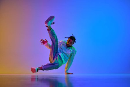 Young guy with dreads, in casual clothes dancing hip-hop, breakdance against gradient multicolored studio background in neon light. Concept of street style dance, fashion, youth, hobby, dynamics, ad