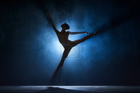 Slim, beautiful, graceful ballerina making creative performance on stage against dark blue background with spotlight. Concept of art, classical ballet, creativity, choreography, beauty, ad
