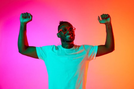 Photo for Win, victory, joy. Strong and handsome african man in glasses looking away over pink-orange background in neon. Concept of sport, musculinity, strenght, healthy lifestyle, hobby, - Royalty Free Image
