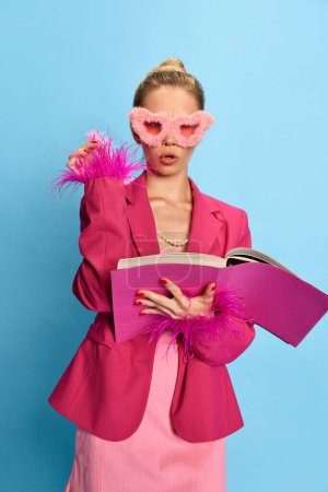 Portrait of cahrming girl, woman wearing glamor in unusual glasses, holding textbook looking at camera on light blue studio background. Concept of fashion, beauty, positive emotions, business.Ad