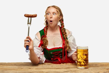 Photo for National cuisine. Surprised young woman dressed traditional dirndl, holding huge juicy,appertizing Bavarian sousage. Concept of Oktoberfest, traditions, drinks and food. Copy space for ad. - Royalty Free Image