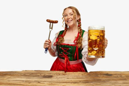 Photo for Smiling young redhead woman dressed traditional dirndl, holding huge juicy,appertizing Bavarian sousage and beer mug. Concept of Oktoberfest, traditions, drinks and food. Copy space for ad. - Royalty Free Image
