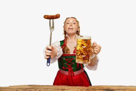 Photo for Young woman dressed traditional dirndl licking lips, serves appetizing Bavarian sausage and beer chole. Concept of Oktoberfest, traditions, drinks and food. Copy space for ad - Royalty Free Image