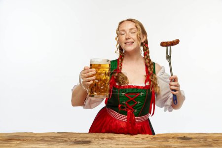 Photo for Waitress. Dreaming redhead woman dressed traditional dirndl, serves appetizing Bavarian sausage on fork and huge beer chole. Concept of Oktoberfest, traditions, drinks and food. Copy space for ad - Royalty Free Image