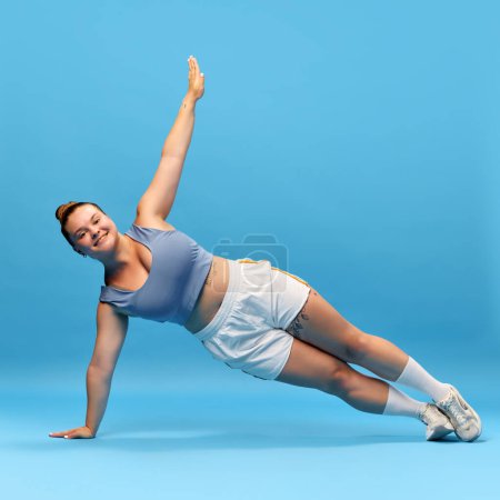 Photo for Young, positive, motivated woman with overweight body doing side plank against blue studio background. Exercising. Concept of sport, body-positivity, weight loss, body and health care - Royalty Free Image