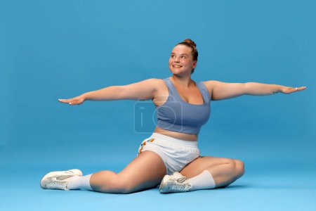 Photo for Self-care and well being. Young overweight woman stretching on floor in sportswear against blue studio background. Motivation. Concept of sport, body-positivity, weight loss, body and health care - Royalty Free Image