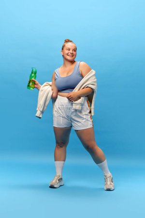 Photo for Full length portrait of young smiling plus-size woman staying in sportswear with bottle for water against blue studio background. rest. energy. Concept of sport, body-positivity, weight loss. - Royalty Free Image