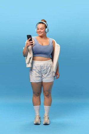 Photo for Full length portrait of young smiling plus-size woman staying in sportswear with phone in headphones against blue studio background. Music for workout Concept of sport, body-positivity, weight loss. - Royalty Free Image