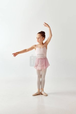 Portrait de petite fille danseuse de ballerine préscolaire adorable en robe de ballet tutu rose pattes blanches debout posant mains levées sur fond blanc. Concept de beauté, mode, passe-temps, expression de soi