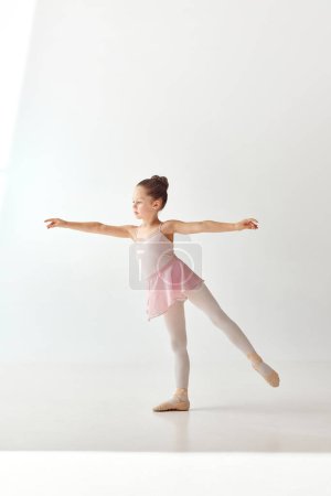Téléchargez les photos : Portrait de petite jolie danseuse de ballerine de chignon d'école en robe de ballet tutu rose, jambes blanches debout posant mains et jambe vers le haut sur fond blanc. Concept de beauté, mode, passe temps. - en image libre de droit