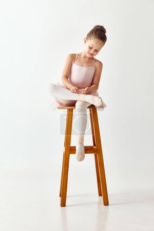 Téléchargez les photos : Portrait de petite danseuse ballerine préscolaire adorable en robe de ballet tutu rose nouant pointe assise sur chaise sur fond de studio blanc. Concept de beauté, mode, passe-temps, performance. - en image libre de droit