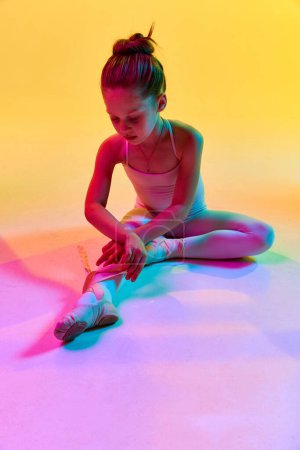 Téléchargez les photos : Portrait de petite danseuse de ballerine préscolaire adorable attachant pointe assise sur le sol dans la lumière au néon mixte sur fond de studio.. Concept d'art, beauté, mode, passe-temps, expression de soi. Publicité - en image libre de droit