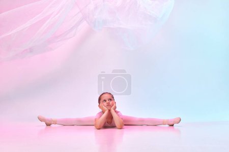 Téléchargez les photos : Portrait de petite fille danseuse de ballerine préscolaire mignonne assise sur le sol dans la pose de ficelle avec tissu volant dans la lumière au néon mixte sur fond de studio. Concept d'art, passe-temps, expression de soi, ballet - en image libre de droit