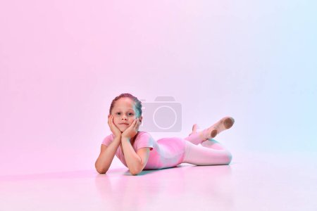 Téléchargez les photos : Portrait de petite danseuse de ballerine mignonne portant un maillot de bain de ballet et pointe couchée sur le sol avec la tête sur les mains dans la lumière au néon mixte. Concept d'art, passe-temps, expression de soi, ballet pour enfants. Publicité - en image libre de droit
