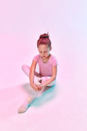Téléchargez les photos : Portrait de petite danseuse de ballerine préscolaire adorable attachant pointe assise sur le sol dans la lumière au néon mixte sur fond de studio. Vue d'en haut. Concept d'art, beauté, mode, passe-temps. Publicité - en image libre de droit