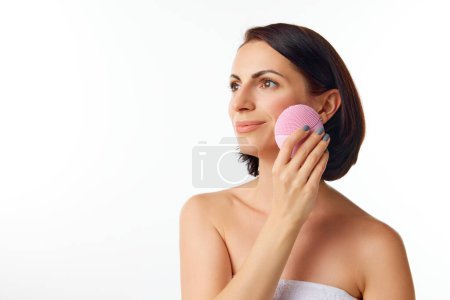 Photo for Young beautiful woman with brunette hair and perfect skin doing skin care procedures with deeply cleansing brush over white color studio background. Beauty routine, cosmetology, youth concept - Royalty Free Image