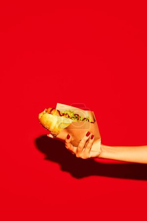 Photo for Hand holding hot delicious traditional American hotdog with beef sausage, sauces with green salad against vivid red background. Concept of fast food, street food, delivery. Copy space for ad, text - Royalty Free Image