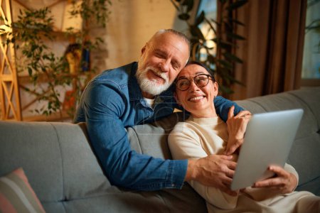 Photo for Portrait of cute and happy mature couple, senior man and woman hugging sitting on sofa with tablet in living room at home. Concept of love, retirement life, pensioners, cozy, winter holidays. - Royalty Free Image