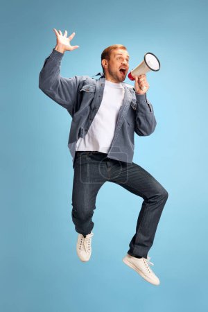 Photo for Portrait of young man jumping of joy shouting to loudspeaker isolated blue studio background. Irritated young guy in casual outfit. Concept of facial expression, emotions, sales, shopping. Ad - Royalty Free Image