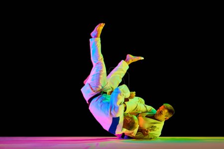 Photo for Athletic men, professional sportsmen during match. Judo fighter attacking his opponent with leg technique in neon light isolated black background. Concept of martial art, combat sport, energy, fit - Royalty Free Image