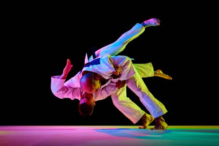 Photo for Portrait of two male sportsmen, martial arts fighters in white kimono fighting performing techniques in motion in neon light isolated black background. Concept of combat sport, energy. Copy space - Royalty Free Image