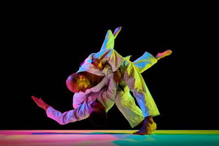 Photo for Two male sportsmen, martial arts fighters in white kimono fighting performing techniques in motion in neon light isolated black background. Concept of combat sport, energy, fit. Copy space, ad - Royalty Free Image