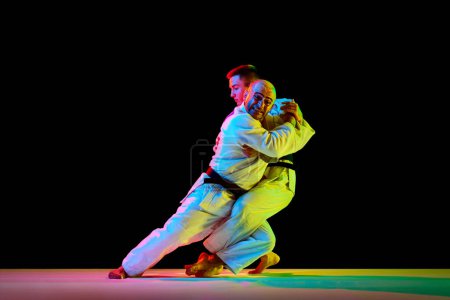 Photo for Two young professional karate men fighting, performing technical skill in neon light isolated black background. Concept of martial art, combat sport, health, strength, energy, fit. Copy space - Royalty Free Image