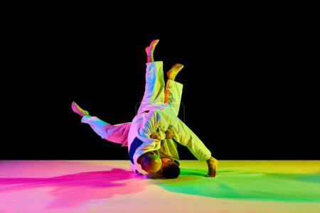 Photo for Two young, athletic professional karate men fighting, performing technical skill in neon light isolated black background. Concept of martial art, combat sport, health, strength, energy. Copy space - Royalty Free Image