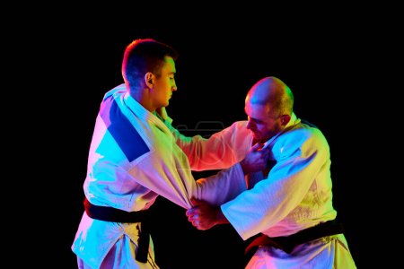 Téléchargez les photos : Portrait latéral de deux hommes, karaté professionnel, combattants de judo en kimono avec ceinture noire démontrent des compétences en lumière au néon isolé fond noir. Concept d'art martial, sport de combat, énergie. - en image libre de droit