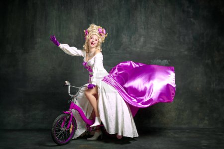 Foto de Mujer encantadora alegre en anticuado, vestido aristocrático sentado, montar en bicicleta para niños contra fondo vintage. Concepto de moda, estilo, vintage, remake, comparación de épocas, hobby, alegría, diversión - Imagen libre de derechos