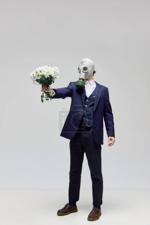 Photo for Attachment defense. Man in black suit and gas mask holds bunch of cute white flowers against white studio background. Allergy to love and feelings. Concept of love, modern relationship, Valentines day - Royalty Free Image