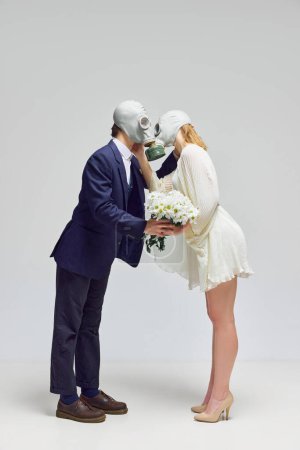 Photo for Fake love, marriage. Man in black suit and woman in white dress holds flowers stands in gas masks and kisses against white studio background. Concept of modern relationship, Valentines day, fake feels - Royalty Free Image