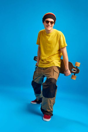 Photo for Full-length portrait of attractive cool attitude in reggae, rasta man style with skateboard against blue studio background. Concept of youth, human emotions, self-expression, subcultures. - Royalty Free Image