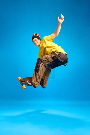 Photo for Positive, smiling man wearing in rastaman, reggie style doing Ollie trick in motion against blue background. Concept of youth, human emotions, self-expression, subcultures, hobby. Ad - Royalty Free Image