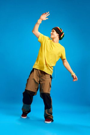 Photo for Happy, cheerful, positive young man in reggae style, rastaman cloth waves friendly against blue studio background. Concept of youth, human emotions, self-expression, subcultures, hobby. - Royalty Free Image