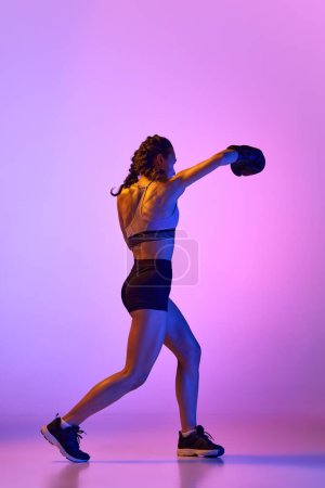 Téléchargez les photos : Image saisissante de femme, sportive professionnelle, boxeuse mettant en valeur ses prouesses en équipement de boxe contre fond de studio dégradé dans filtre au néon. Concept de sport, mode de vie actif, santé, compétition - en image libre de droit