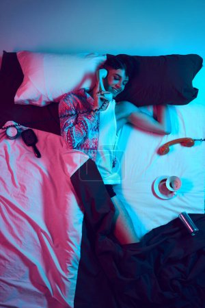 Photo for Gender fusion symphony. Visual overture. Creative portrait of young male model lying in bad and talking on retro phone as woman in neon light. Concept of self-expression, masculinity and femininity. - Royalty Free Image