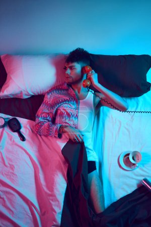 Photo for Studio photo of man with make up on one half of face and dressed as man and woman lying in bed talking on retro phone in neon light. Concept of self-expression, masculinity and femininity, diversity. - Royalty Free Image
