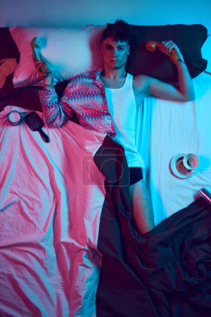 Photo for Connection. Bridging Masculine and Feminine Energies. Young male model lying in bed and holding landline retro phone and looking at camera in neon light. Concept of lifestyle, masculinity, femininity. - Royalty Free Image