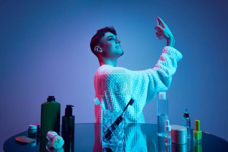 Photo for Beyond Binary Beauty. Male model, whose face reveals both its masculine and feminine sides amidst neon-lit bathroom, surrounded by cosmetic products. Concept of beauty and fashion, self-care. Ad - Royalty Free Image