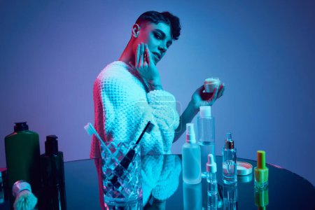 Photo for Unveiling Radiance. Masculinity meets femininity, Male model surrounded by cosmetic products in neon-lit bathroom. Journey of self-care and beauty. Concept of gender equality, body care. Ad - Royalty Free Image
