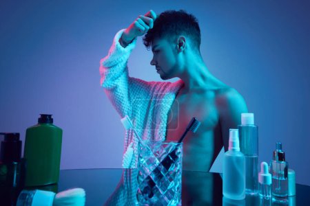 Photo for Dynamic elegance of young man portraying both masculine strength and feminine grace in neon-lit bathroom. Concept of beauty and fashion, self-care and cosmetic allure, health, lifestyle. Ad - Royalty Free Image