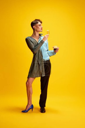 Photo for Young male model half-wearing in womans and mans clothes posing holding glasses of champagne and whiskey in hands on his female and male sides. Concept of self-expression, fashion and style, beauty. - Royalty Free Image