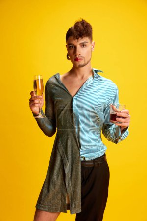 Photo for Portrait of person half-wearing in womans and mans clothes posing holding glasses of champagne and whiskey in hands on his female and male sides. Concept of self-expression, fashion and style. Ad - Royalty Free Image