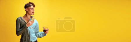 Photo for Banner. Soft and liquor drinks. Male model with make up man and woman holding glasses of champagne and whiskey against yellow background with copy space. Concept of self-expression, lifestyle. Ad - Royalty Free Image