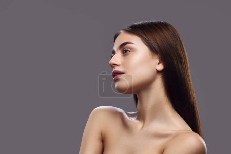 Profil latéral d'une jeune femme aux cheveux lisses et aux épaules nues, présentant un look propre et élégant sur fond gris. Concept de soins capillaires, cheveux forts, sains et brillance naturelle. Publicité