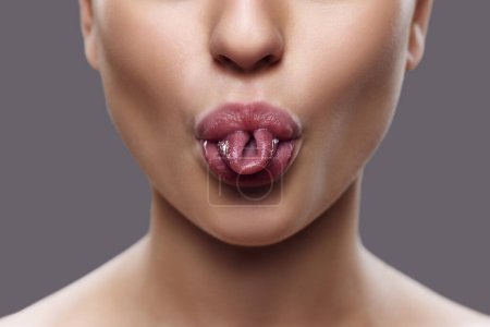 Ausgeschnittenes Foto von. Großaufnahme von Frauenmund mit verdrehter Zunge, unterstrichen durch subtilen Lipgloss, vor grauem Hintergrund. Konzept der Übungen für Gesichtsmuskeln, Augmentation, Verjüngungskur