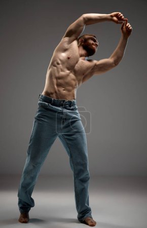 Photo for Shirtless, fit male stretching arms overhead posing blue jeans against grey studio backdrop. Movement and flexibility. Concept of beauty and fashion, masculinity, bodybuilding, style. - Royalty Free Image