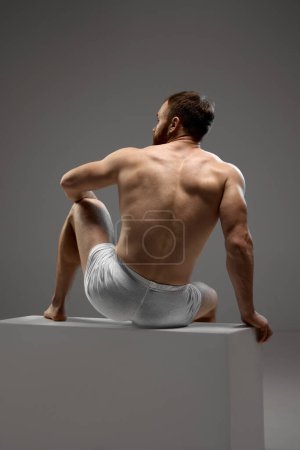 Photo for Rear view of muscular man, model seated on white stage in white underwear against grey studio background. Concept of beauty and fashion, masculinity, bodybuilding, style. - Royalty Free Image